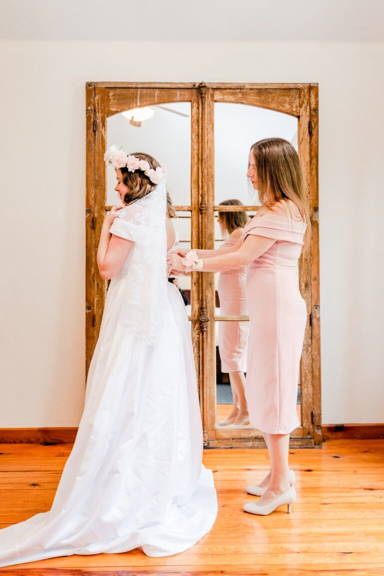 Mother helping her daughter with her wedding dress in front of a mirror, illustrating mother-of-the-bride attire guidance