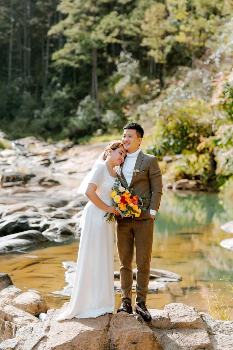 Couple at eco-friendly wedding near a forested stream holding vibrant bouquet – sustainable wedding concept.